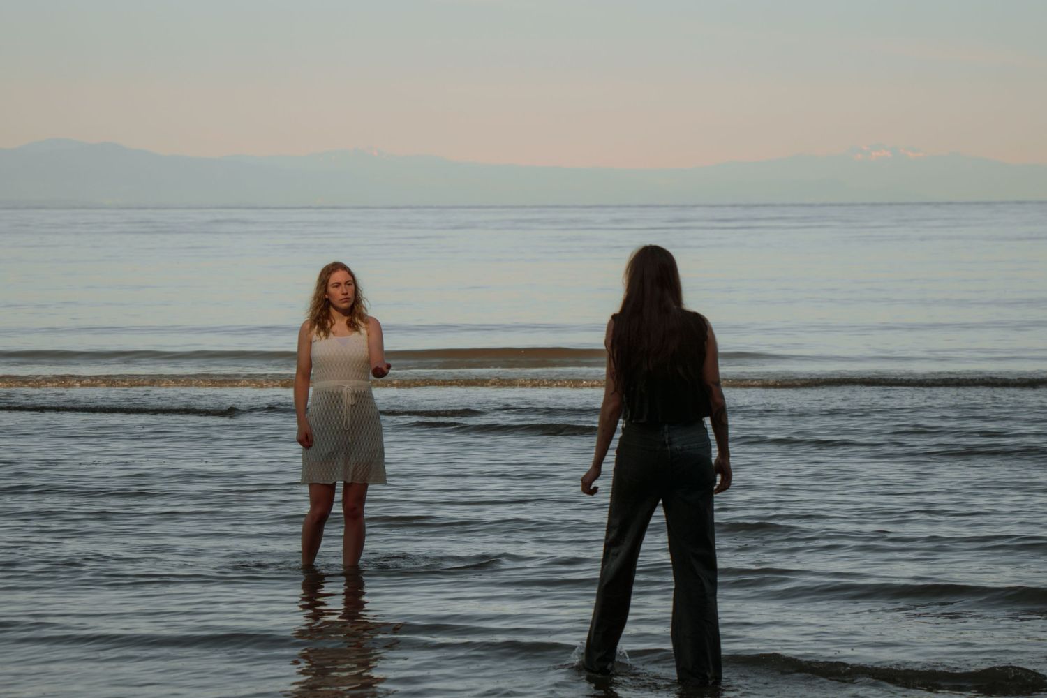Two figures standing in shallow ocean water during twilight with mountains in the background.