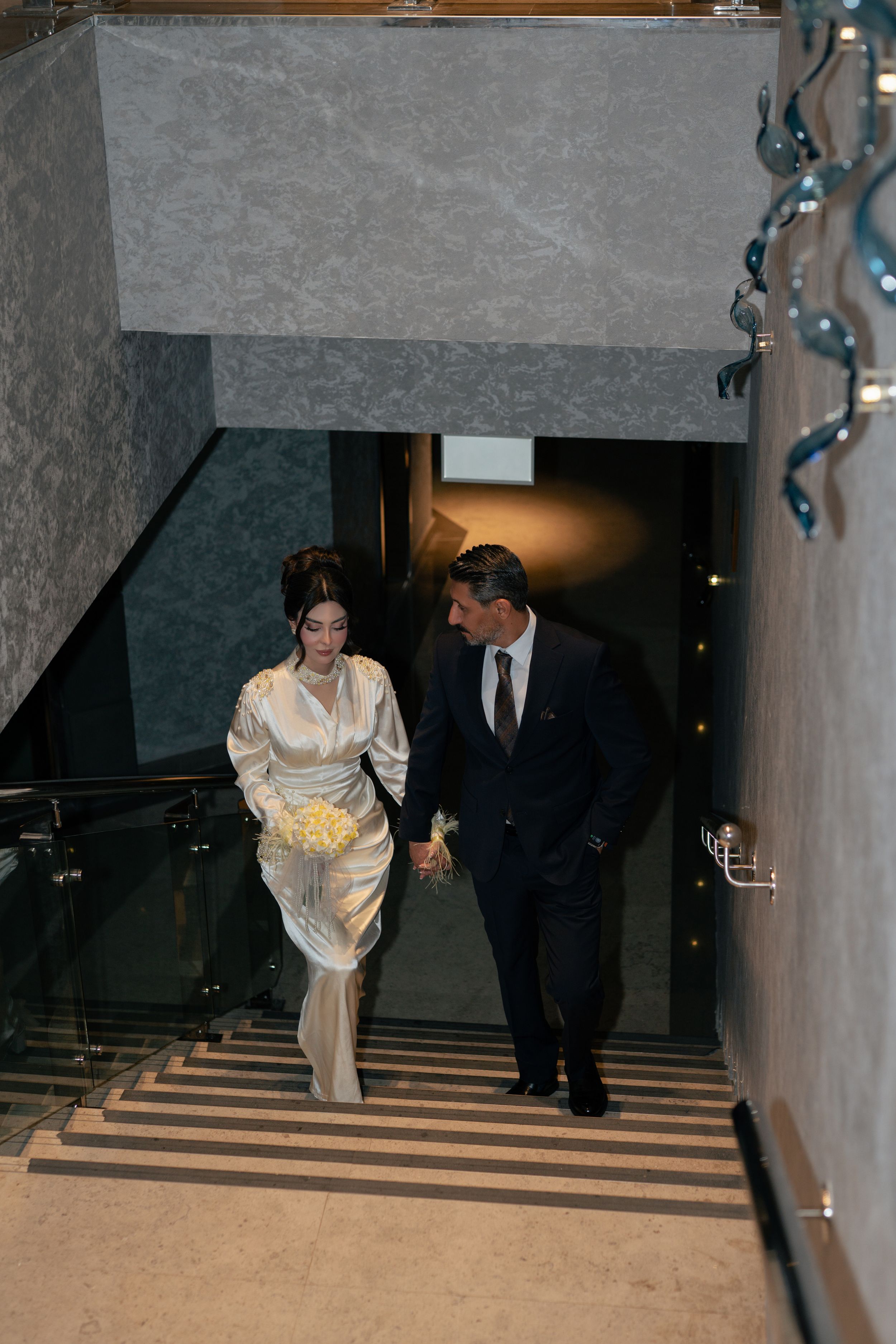 A couple descending a grand staircase at an elegant event, with the woman wearing a light-colored formal gown.