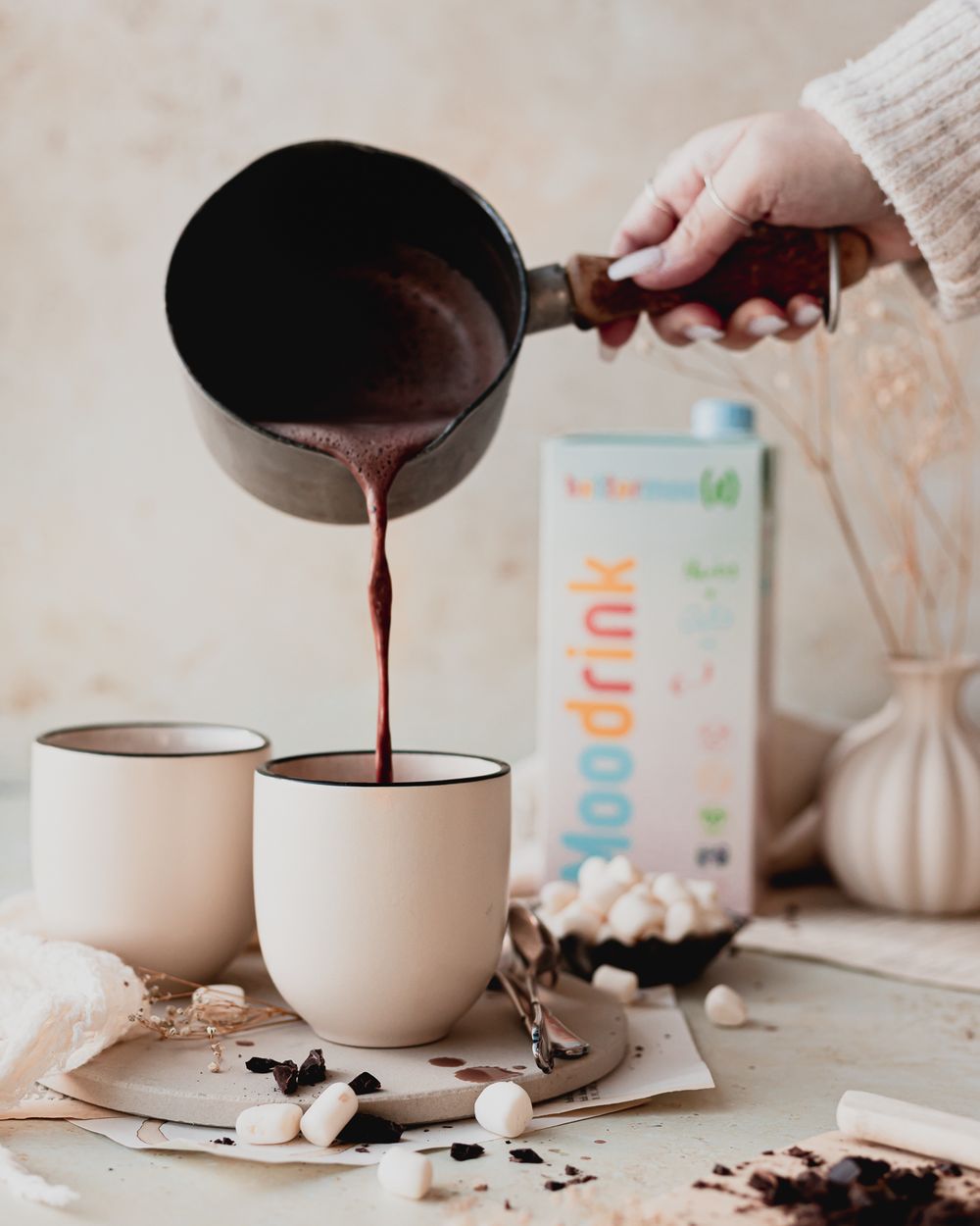 A hand pours hot chocolate from a rustic saucepan into a mug. Nearby are mini marshmallows and chocolate shavings.