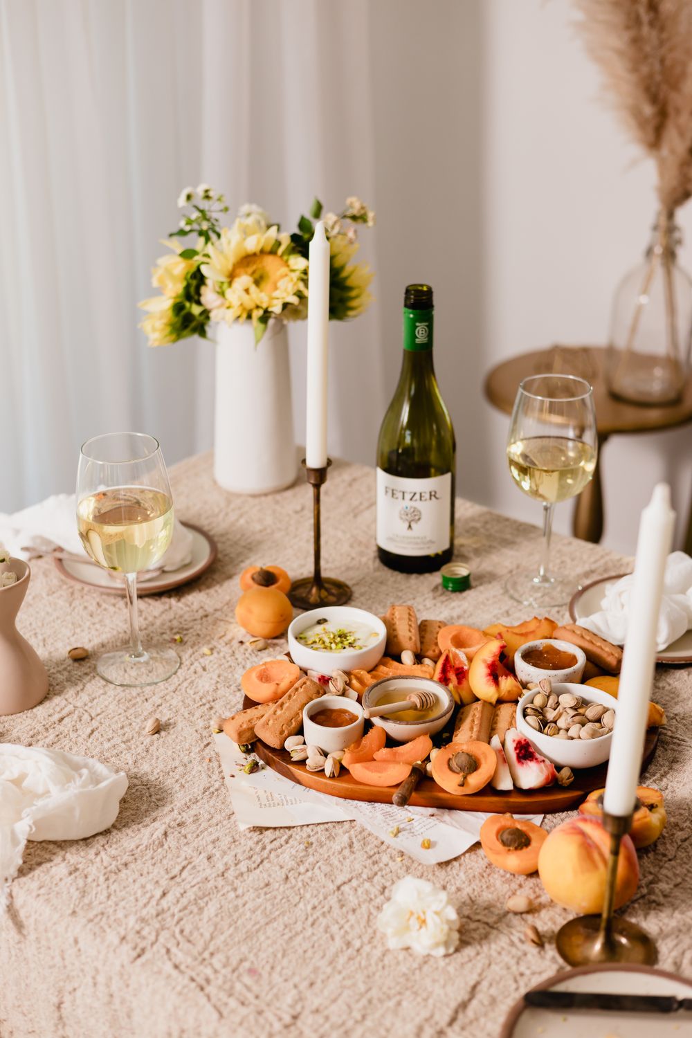 A table set for two with wine, candles, and a platter of fruits, nuts, and dips on a textured cloth. A vase with yellow