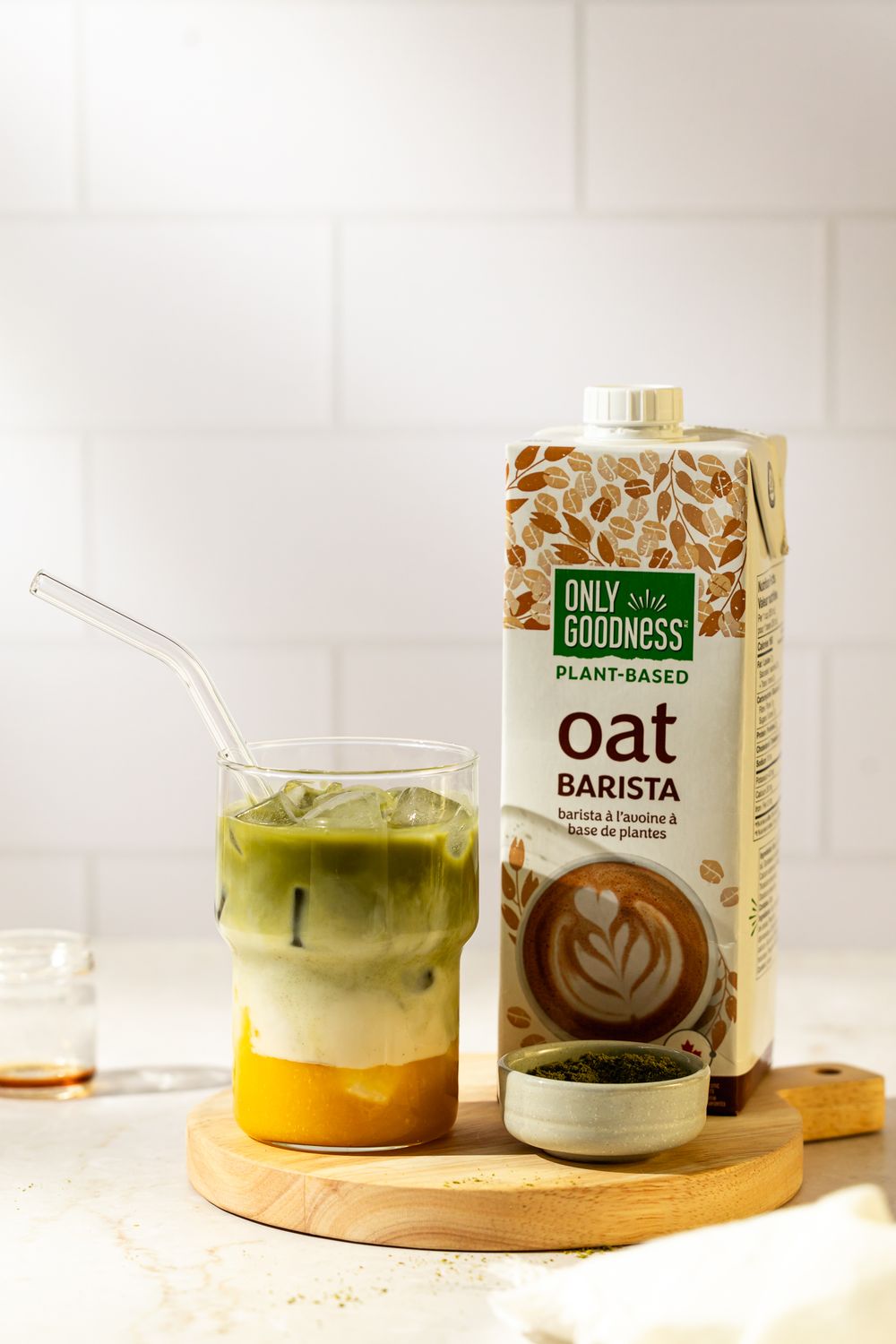 Layered mango matcha latte with oat milk and ice, styled with matcha powder and carton on a wooden board in soft light.