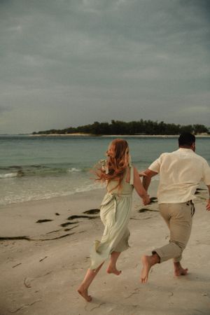 A couple runs barefoot on a scenic beach with turquoise water and a tropical island in the background at sunset.