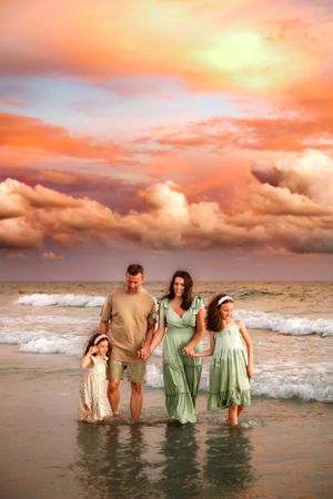 A family stands together in the ocean at sunset with dramatic orange and pink clouds in the sky above.