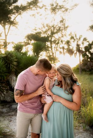 A couple shares a tender moment with their baby at sunset near tropical foliage and water.