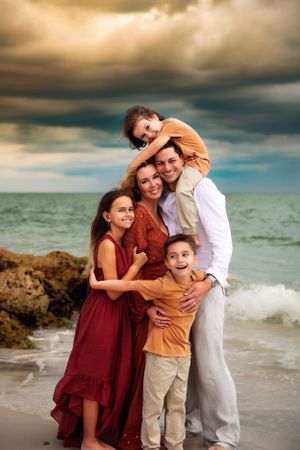 A family poses together on a beach at sunset with dramatic stormy clouds and ocean waves in the background.