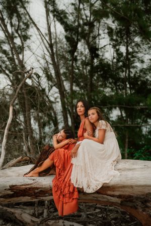 Two women in flowy dresses sit together on a fallen tree trunk at dusk with greenery in the background.