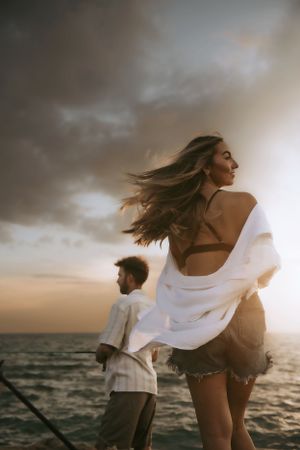 Couple enjoying sunset by the ocean with wind blowing through their hair and white flowing fabric in golden light.