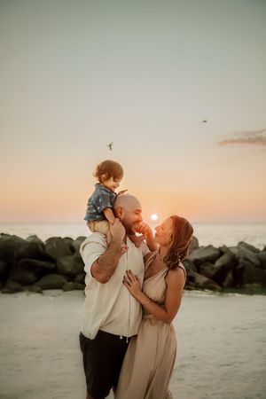 Family of three enjoying a golden sunset on the beach, father holding toddler on shoulders while mother smiles lovingly.