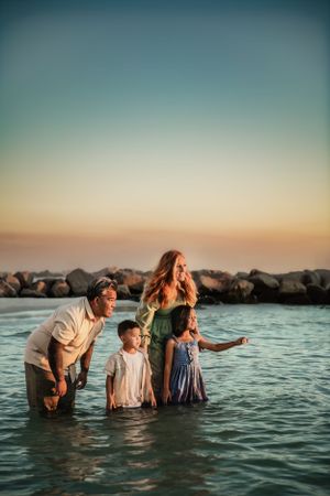 Family enjoying a sunset beach moment standing together in shallow turquoise water with rocky coast in background.