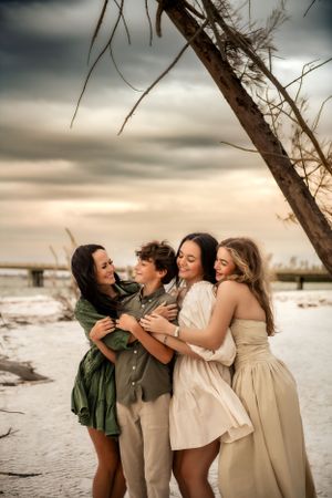 A group of friends embrace and laugh together on a beach at sunset while wearing flowy dresses in neutral tones.