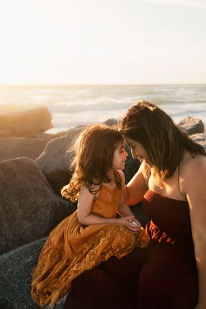 A tender moment shared between mother and daughter in flowing dresses on coastal rocks at golden hour sunset.