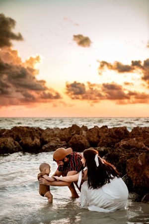 A couple in casual beach attire plays with their baby in shallow ocean water at sunset while rocky cliffs frame the scene.