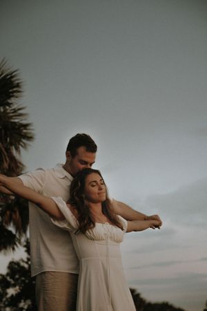 A couple embraces outdoors at dusk, with the woman in a white dress stretching her arms against a moody sky and palm trees.