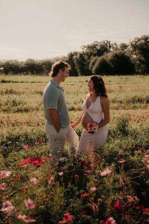 Couple standing in a blooming wildflower field at golden hour, woman in white dress holding bouquet, man in light blue polo.
