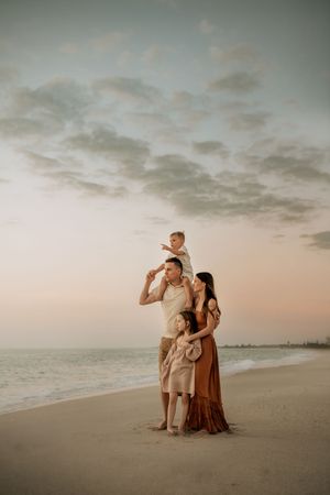 Family of four poses on a sandy beach at sunset, with child on father's shoulders, warm earth tones.