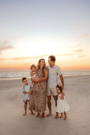 A family of five stands together on a sandy beach at sunset with a warm glowing sky in the background.