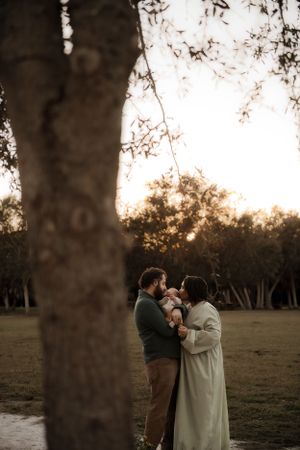 A scenic sunset moment captured between a couple holding an infant beneath a tree in a park setting.