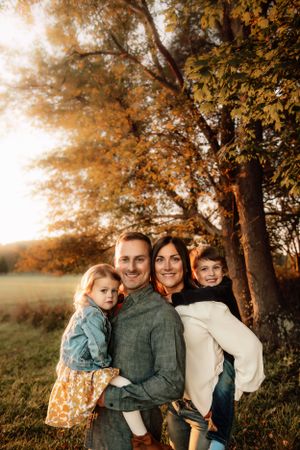 A family stands together outdoors in autumn, with golden sunlight filtering through trees in the background.