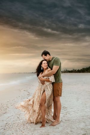 A romantic couple embraces on a beach at sunset with dramatic storm clouds overhead.
