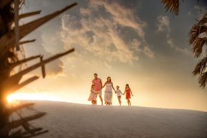 Family walking hand-in-hand on a sandy beach at golden sunset with palm trees and dramatic clouds.
