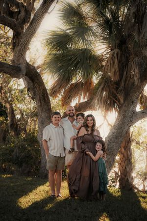 Family of five posing under palm trees in golden hour light, mother in brown gown, outdoors.