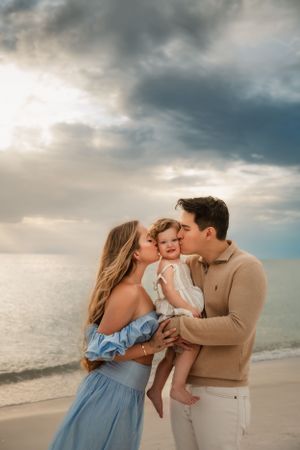 A couple shares tender kisses with a baby at sunset on a beach with dramatic cloudy skies in the background.