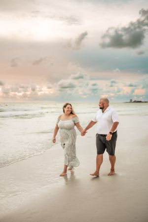 A couple walks hand in hand along a serene beach at sunset with cloudy skies overhead.