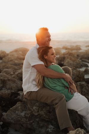 A couple embraces while sitting on coastal rocks during golden hour sunset, creating a romantic silhouette against the ocean.