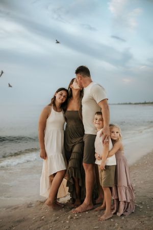 A family stands together barefoot on a beach at dusk with seabirds flying overhead.