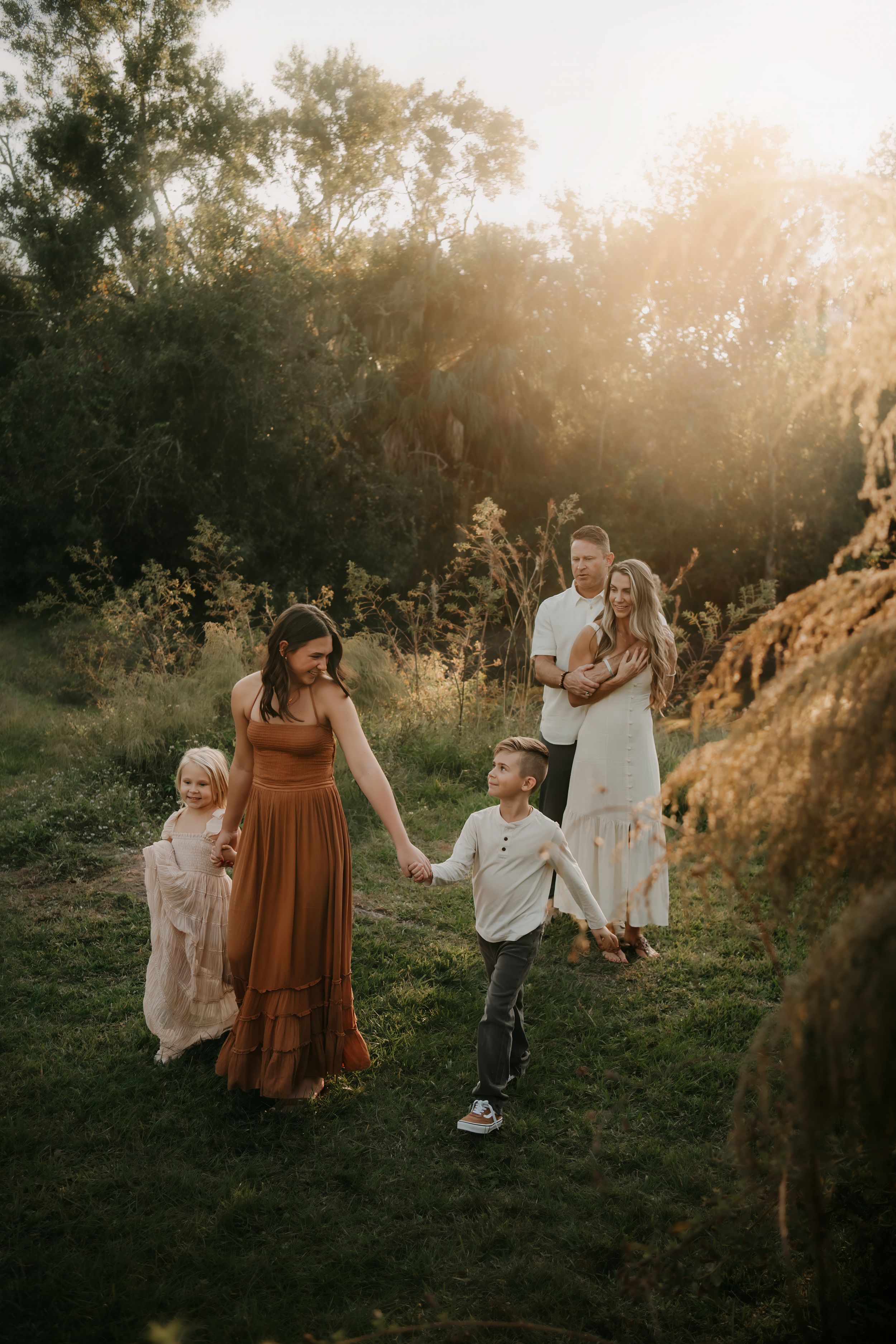 Family of four enjoying a golden hour outdoor photo session in a lush green field surrounded by trees.