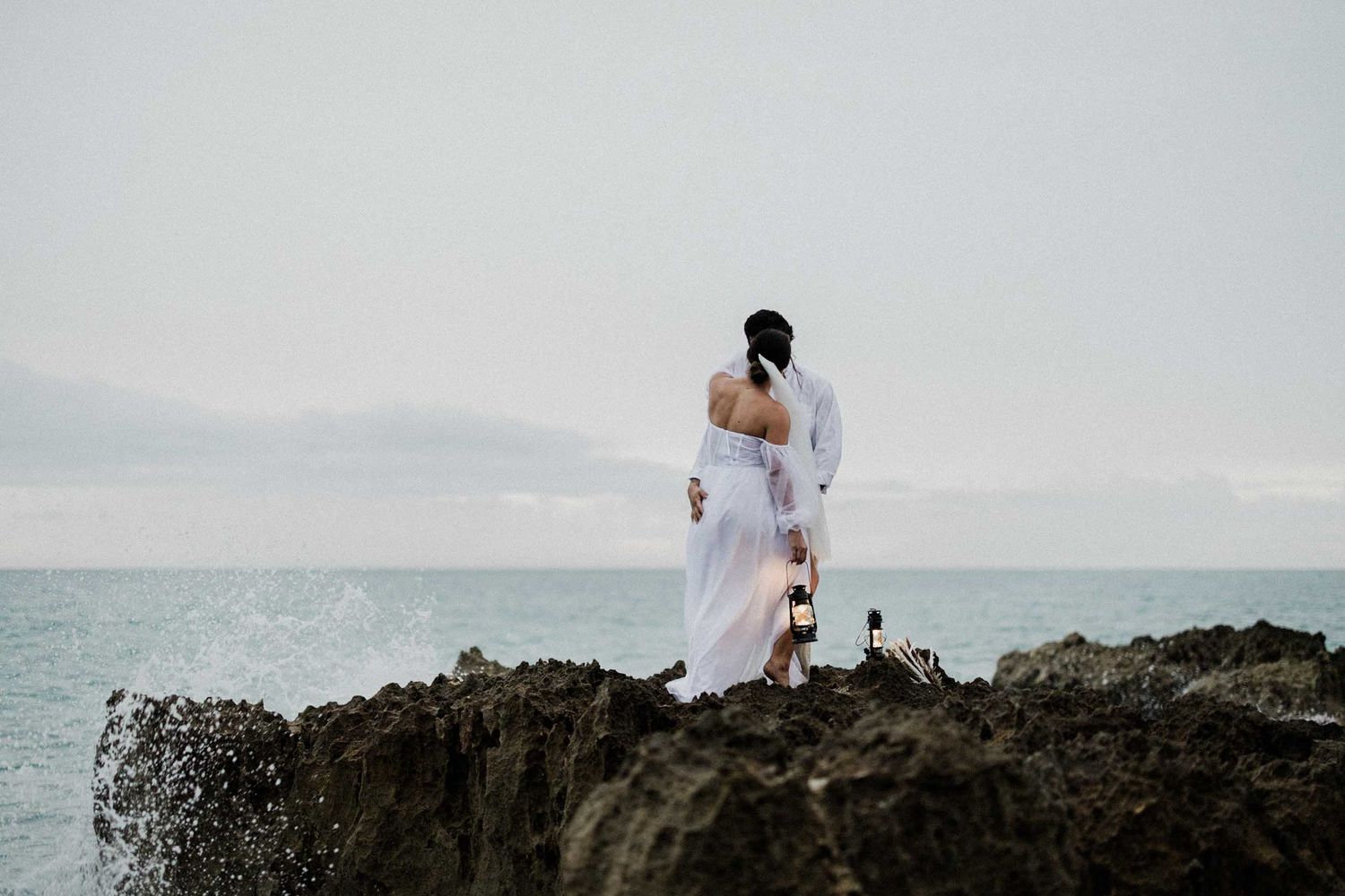 A couple in white attire stands on rocky cliffs overlooking a misty ocean horizon during a romantic photoshoot.