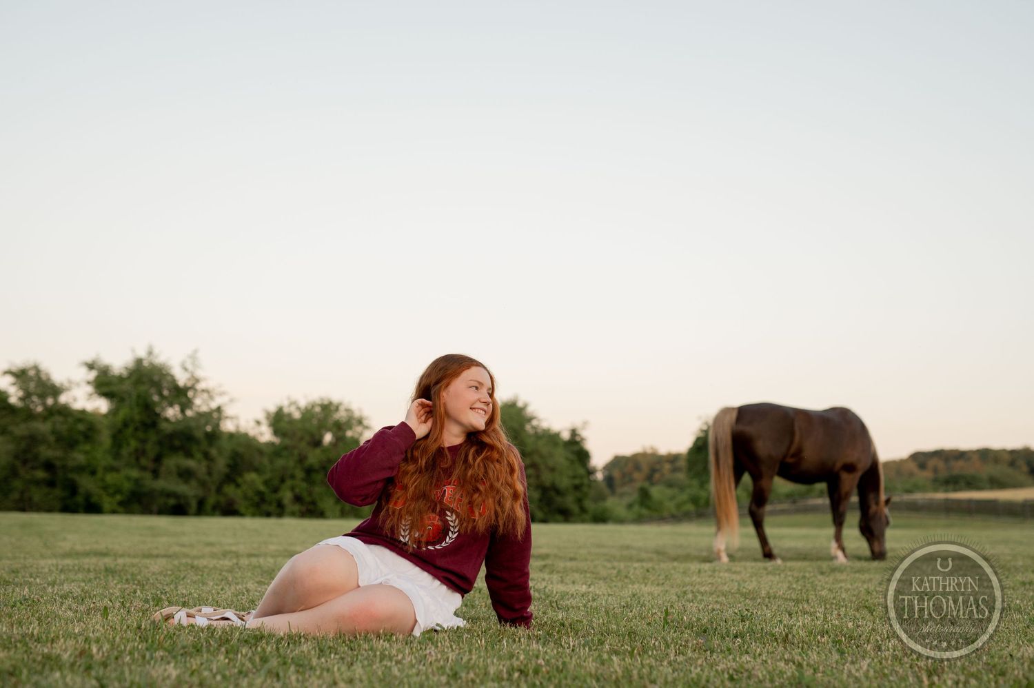 Person rests on grass at sunset while horse grazes in distant field.