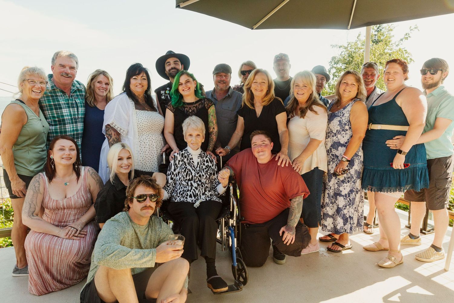 A large group poses together for a casual outdoor gathering under a patio umbrella.