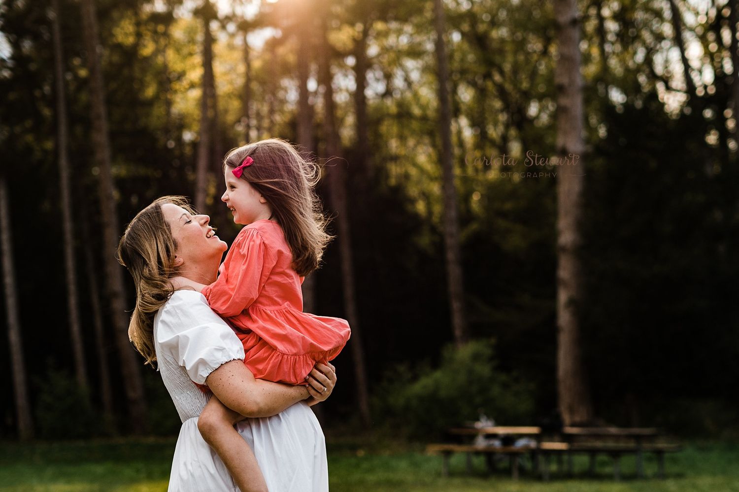 Pregnant mother with white dress, lovingly carrying her daughter with a red dress and looking at her face on a sunny day in Switzerland