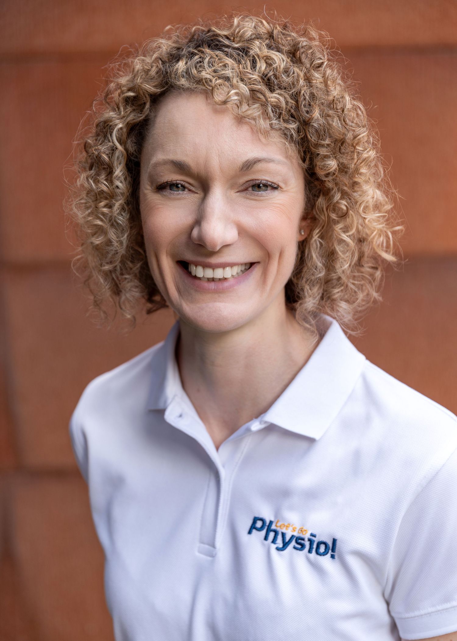 Healthcare professional wearing a white Physiol polo shirt smiling warmly against a brown background.