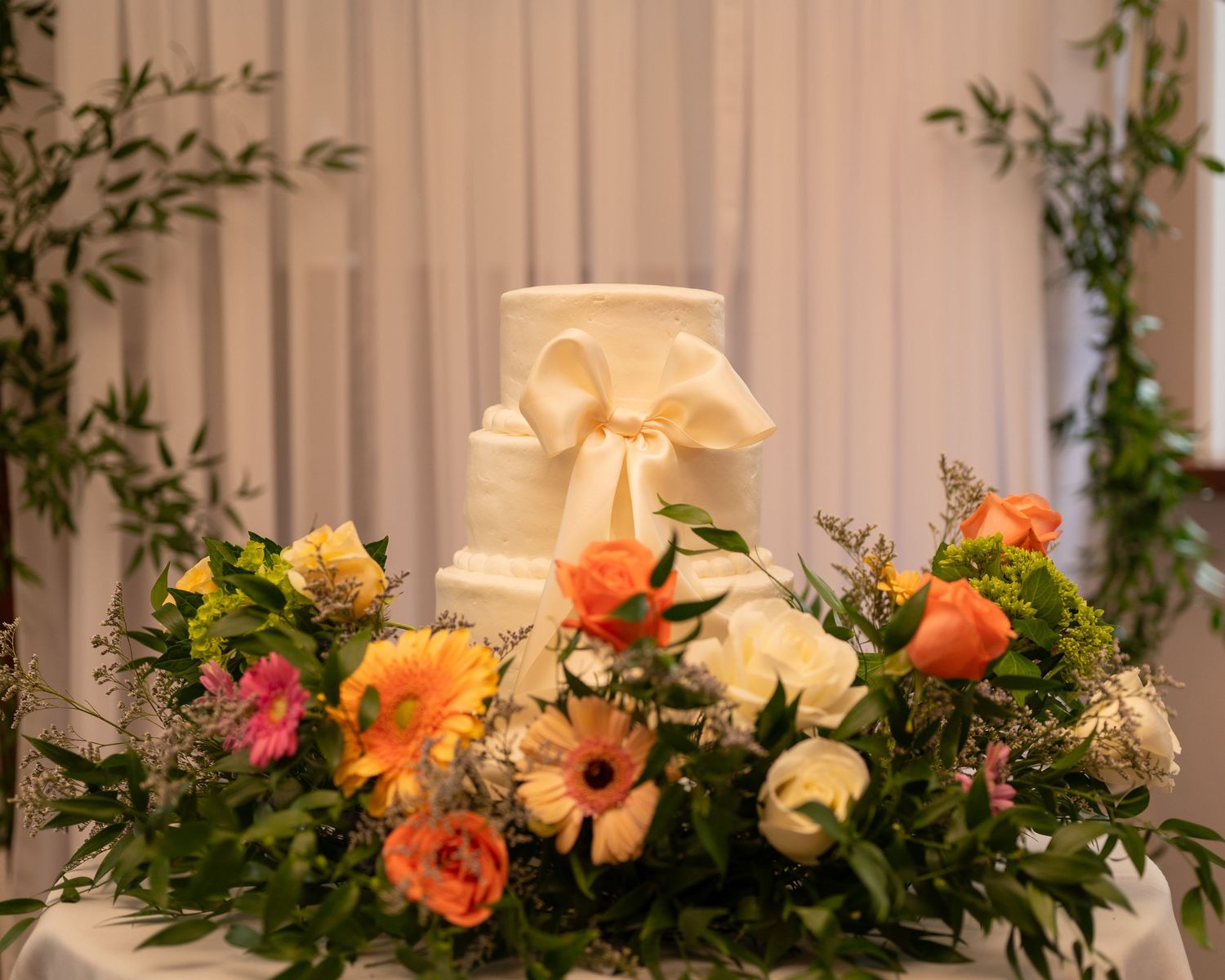 Elegant white wedding cake adorned with peach bow and surrounded by vibrant coral and yellow floral arrangements.