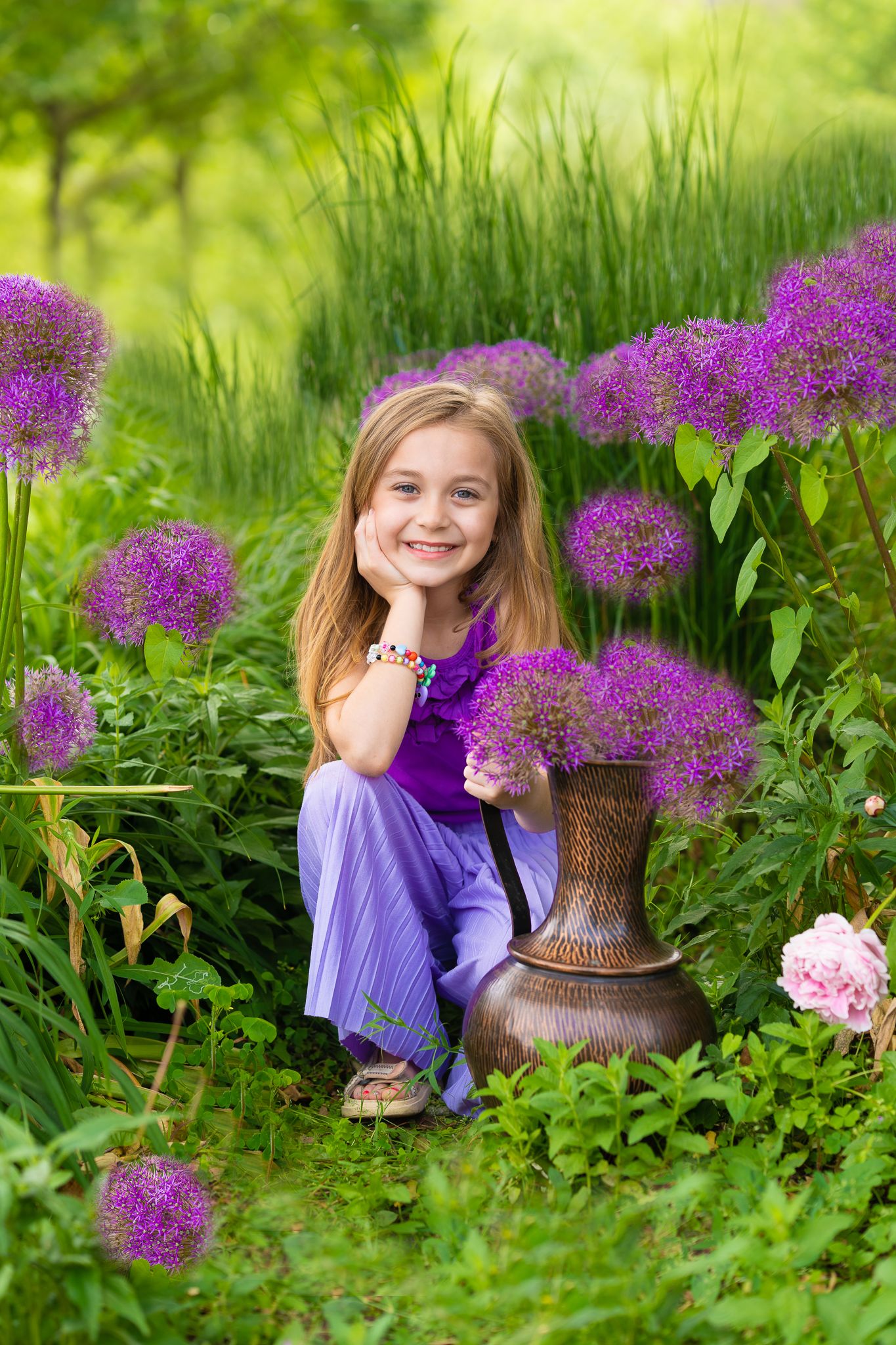 Kinderfotografie - Young girl in purple dress poses with vibrant allium flowers in a lush summer garden.