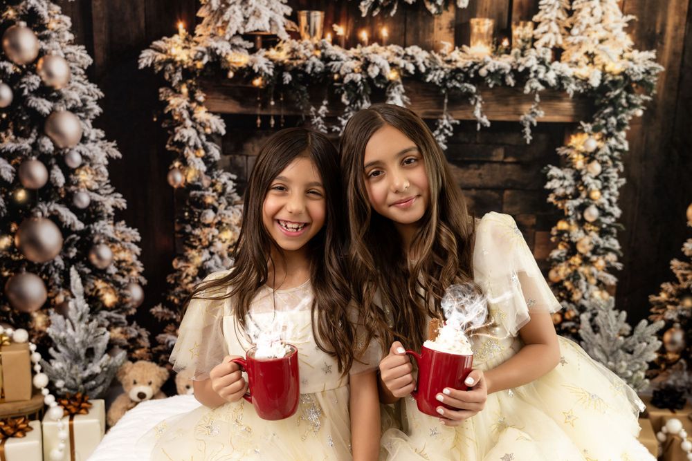 Two people in white dresses hold red beverages amid snowy Christmas decorations and twinkling lights.