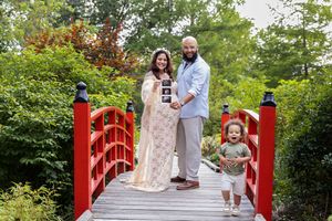 Family poses for photos on a red wooden bridge surrounded by lush green trees in a garden setting.