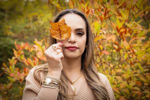 Person holding an autumn leaf in front of their face while wearing a beige sweater against colorful fall foliage.