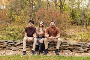 Three people sitting together on a stone wall surrounded by autumn foliage in a garden setting.