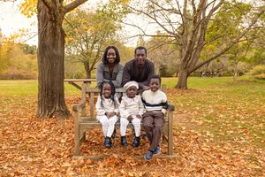 Family sits together on a fallen tree trunk surrounded by colorful autumn leaves in a park setting.