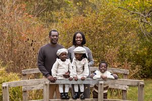 Family wearing coordinated white and brown outfits sits together on wooden bridge during autumn photo session.