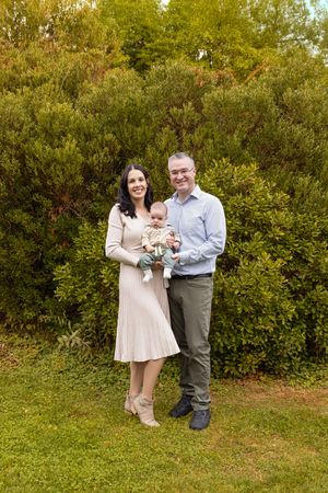 A couple stands together in a lush garden setting during autumn, wearing light colored outfits and embracing.