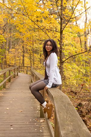 Person in white blazer sits on wooden railing of forest boardwalk with golden autumn leaves in background.