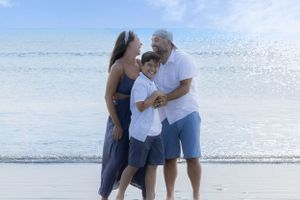 A family of three shares a tender moment together while standing on a sunny beach near the ocean.