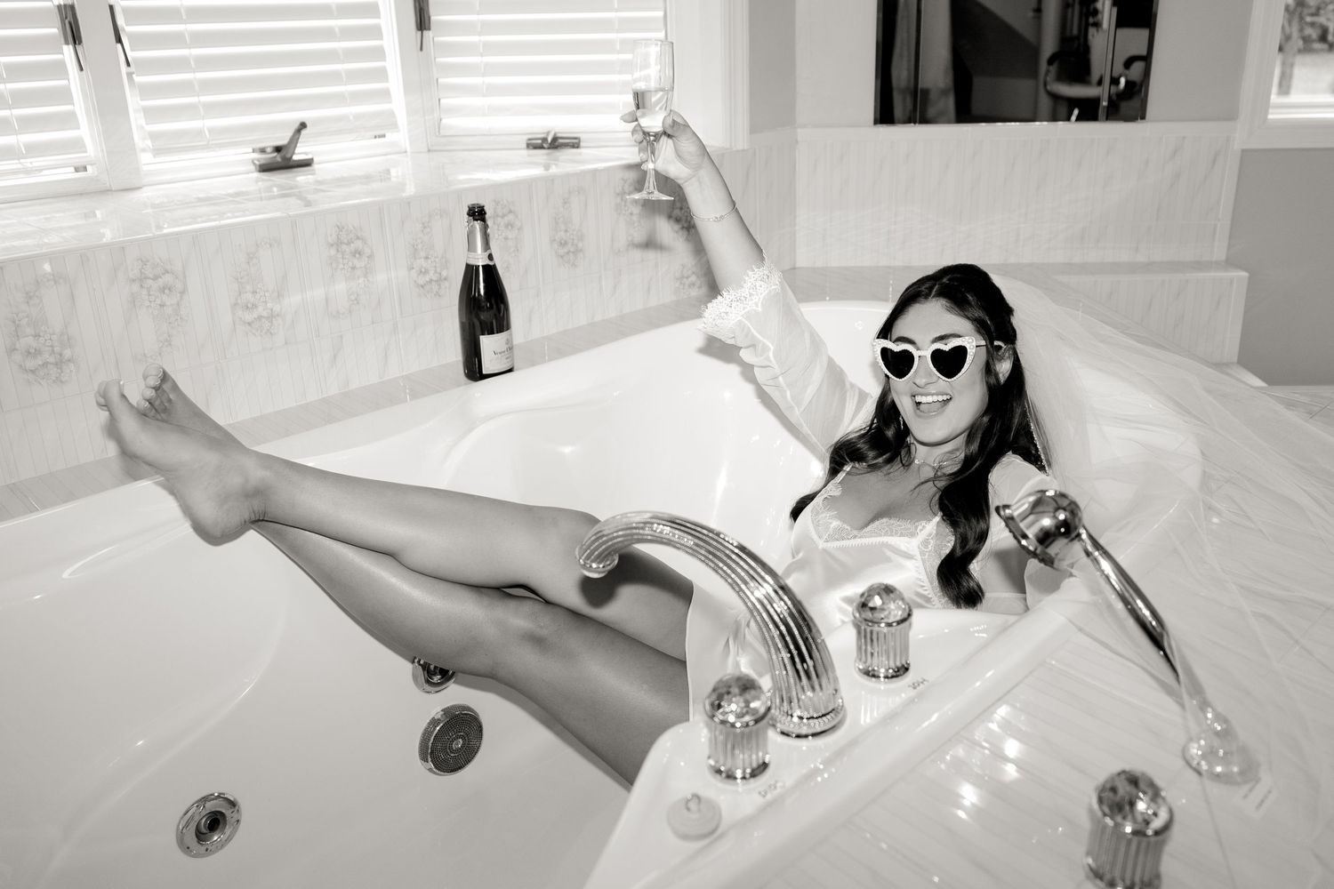 Black and white photo of someone relaxing in a luxurious bathtub with champagne and wearing sunglasses.