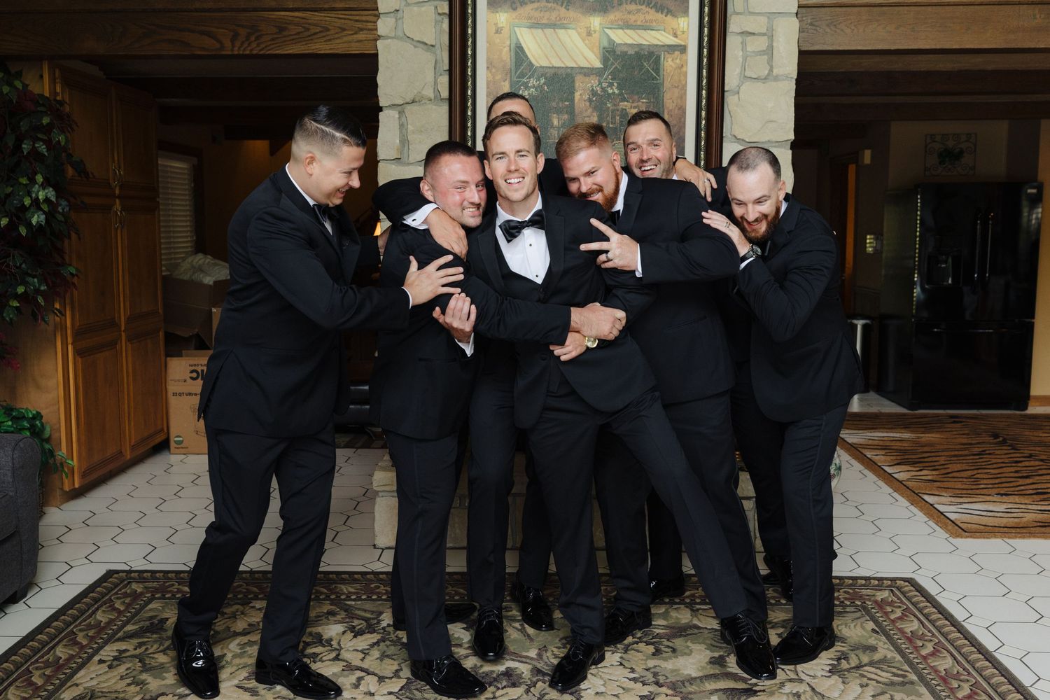 A fun group photo of groomsmen in black suits playfully posing together on a patterned carpet.