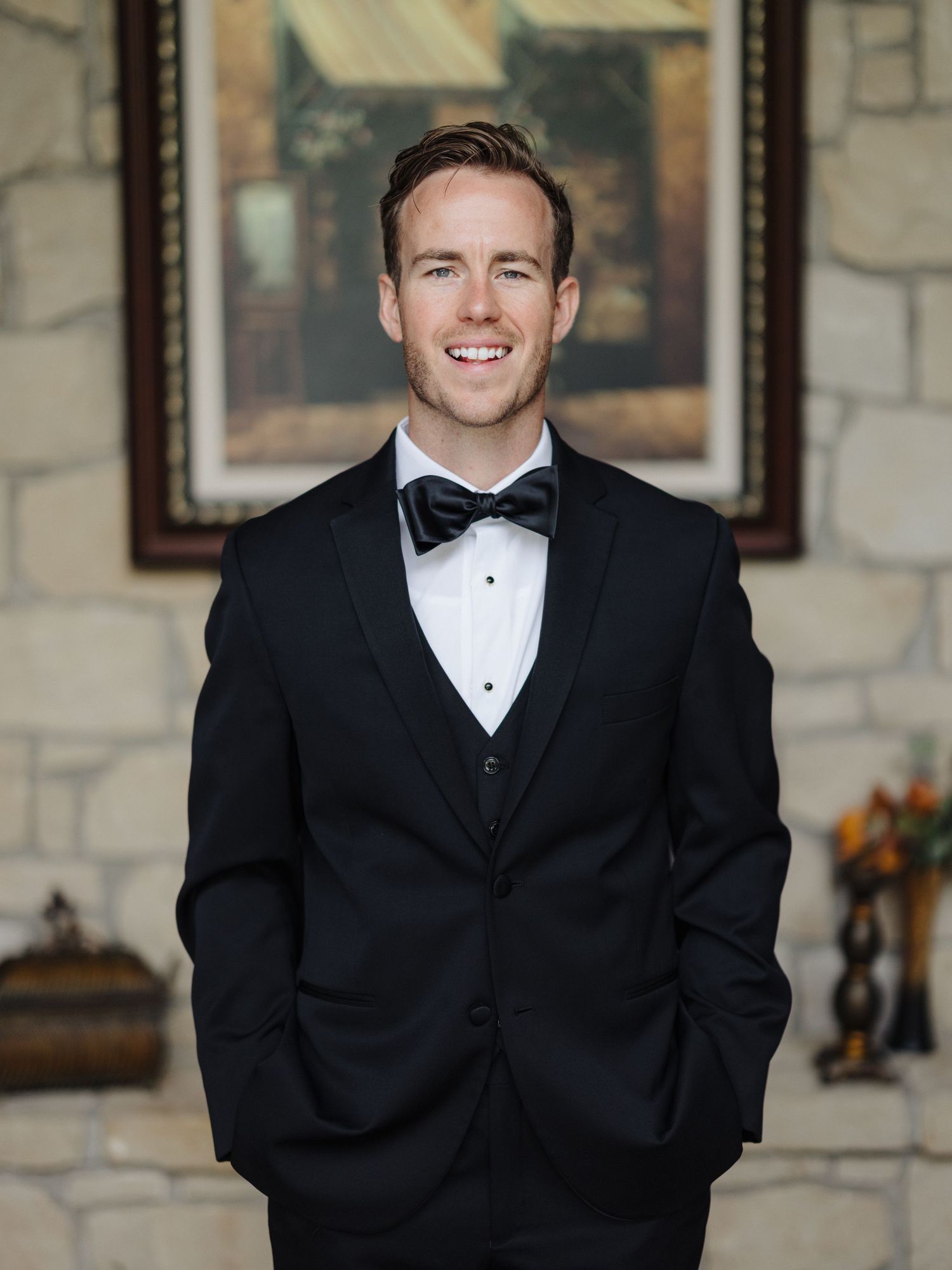 Elegant groom dressed in classic black tuxedo with bow tie stands smiling in formal portrait setting.