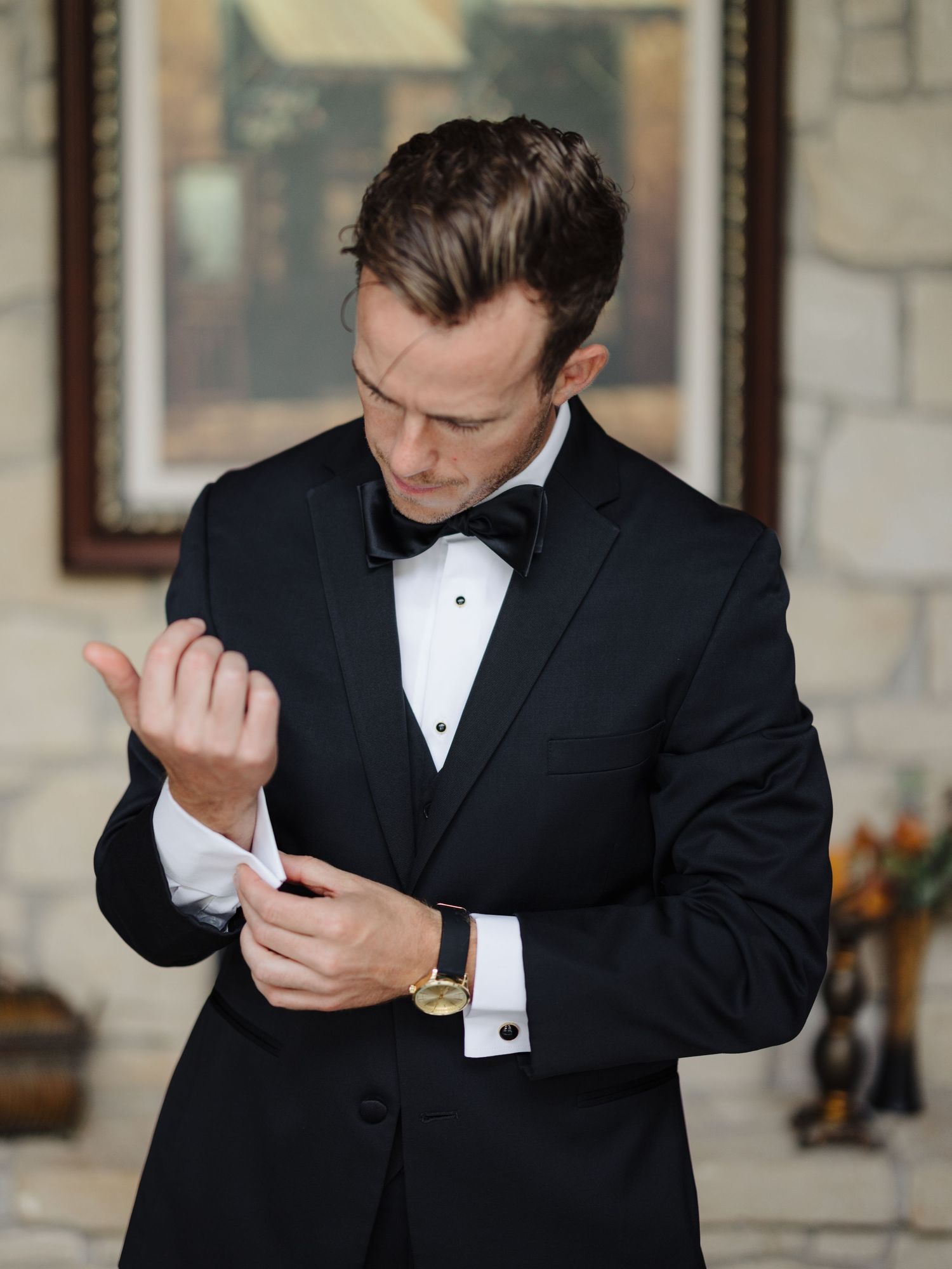 Person in black tuxedo and bowtie adjusts cufflink while getting ready.